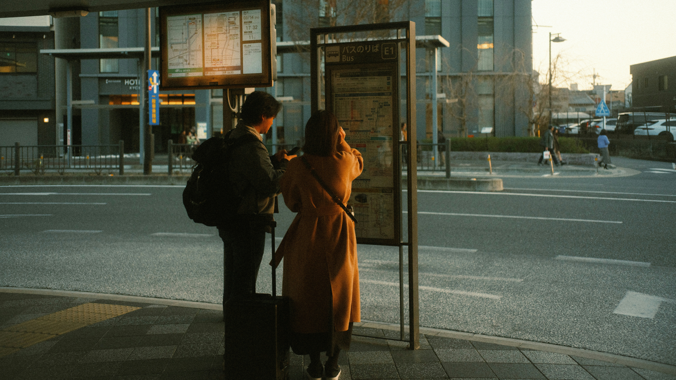 Two people standing by a bus stop in Japan on a city street at winter sunset.