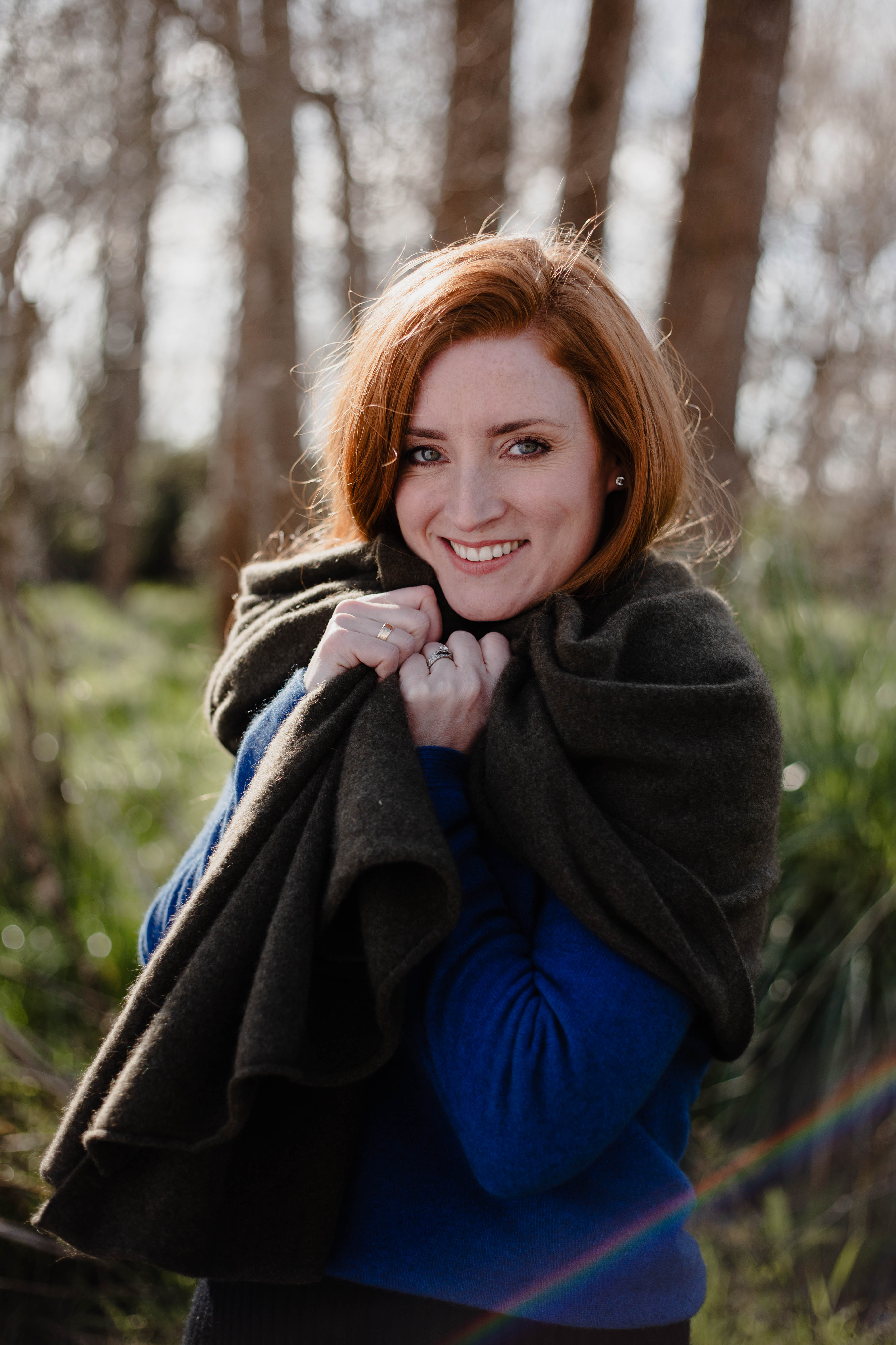 Woman with red hair standing in a forest, wearing a blue sweater and dark coat. She wears New Zealand brand Native World